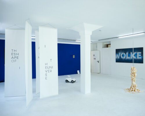 Heller Galerieraum mit blau gestrichener Rückwand. Links hängen mehrere schmale, weiße Textilbanner mit schwarzer, fragmentierter Schrift von der Decke. In der