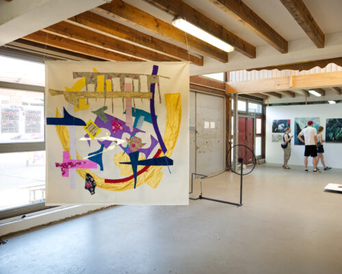 Innenraum einer modernen Kunstgalerie mit Betonboden und sichtbaren Holzbalken an der Decke. Im Vordergrund hängt ein großes, buntes Textilkunstwerk mit abstrak