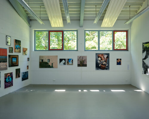 Heller Galerieraum mit weiß gestrichenen Wänden und grauem Boden, an denen zahlreiche farbige Fotografien und Kunstwerke in unterschiedlichen Formaten hängen. L