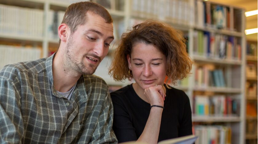 Zwei junge Erwachsene sitzen in einer Bibliothek nebeneinander und lesen gemeinsam in einem Buch, das vor ihnen auf dem Tisch liegt.