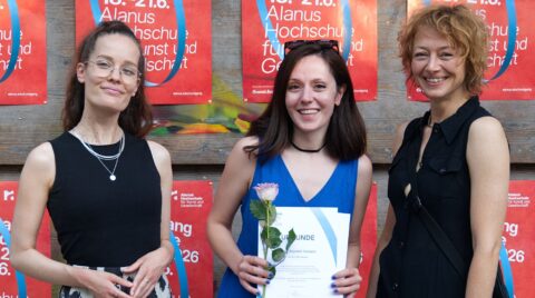 Drei lächelnde Frauen stehen nebeneinander vor roten Plakaten der Alanus Hochschule. Die Frau in der Mitte trägt ein blaues Kleid, hält eine Urkunde und eine ei