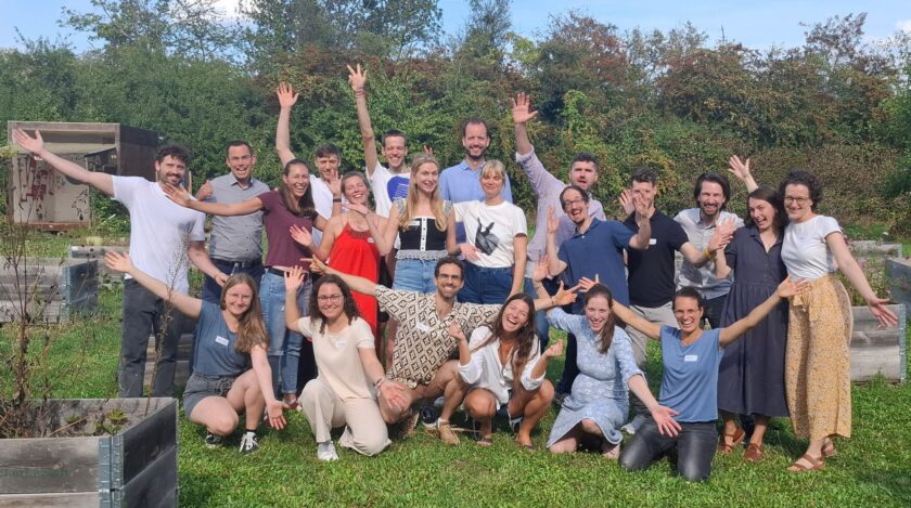 Eine große Gruppe von etwa zwanzig lachenden Menschen unterschiedlichen Alters posiert im Freien auf einer Wiese, viele knien oder hocken vorne, andere stehen d