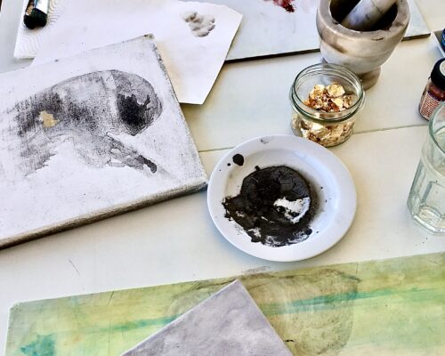 Auf einem Arbeitstisch liegen mehrere Leinwände und Papiere mit grauen und pastellfarbenen Malereien, daneben Pinsel auf Küchenpapier, ein Mörser mit Stößel, ei