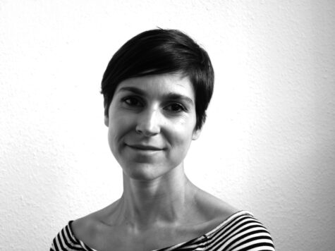 Schwarz-weiß-Porträt einer lächelnden Person mit kurzem, dunklem Haar vor einer hellen, strukturierten Wand, im gestreiften Oberteil.