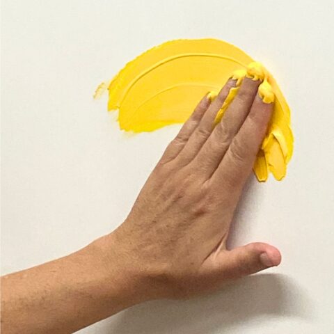 Eine Hand verstreicht mit den Fingern leuchtend gelbe Farbe halbkreisförmig auf einer weißen Fläche.