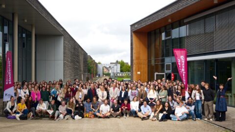 Große Gruppe junger Menschen posiert für ein Gruppenfoto im Innenhof zwischen zwei modernen Hochschulgebäuden. Viele sitzen vorn auf dem Boden oder auf Stufen,