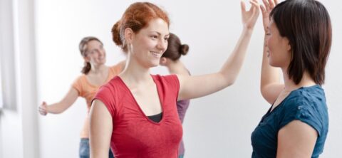 Vier Frauen stehen in einem hellen Raum und üben gemeinsam eine Bewegungs- oder Tanzübung; zwei Frauen im Vordergrund lächeln sich an und berühren einander mit