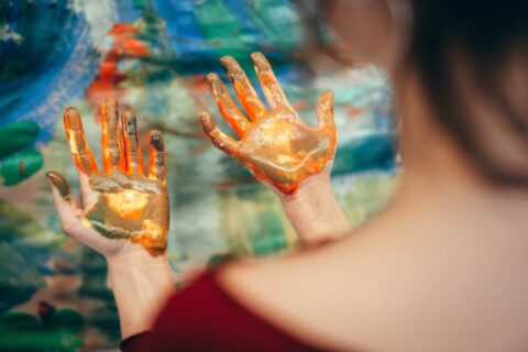Zwei hochgehaltene Hände einer Person, die mit orangefarbener, gelber und goldener Fingerfarbe bedeckt sind, vor einem bunten, unscharfen Hintergrund eines abst